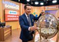Apresentador da Loteria dos Sonhos segura a bola 37 ao lado de um globo transparente em estúdio laranja