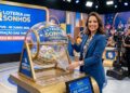 Apresentadora mulher sorrindo ao girar globo de sorteio na Loteria dos Sonhos, estúdio iluminado, público ao fundo