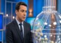 Apresentador de terno azul-marinho ao lado de globo de sorteio com bolas numeradas em estúdio de TV