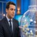 Apresentador de terno azul-marinho ao lado de globo de sorteio com bolas numeradas em estúdio de TV