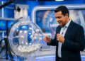 Homem de blazer azul-marinho e camisa clara observa bolas numeradas em globo transparente de sorteio em estúdio de TV