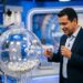 Homem de blazer azul-marinho e camisa clara observa bolas numeradas em globo transparente de sorteio em estúdio de TV
