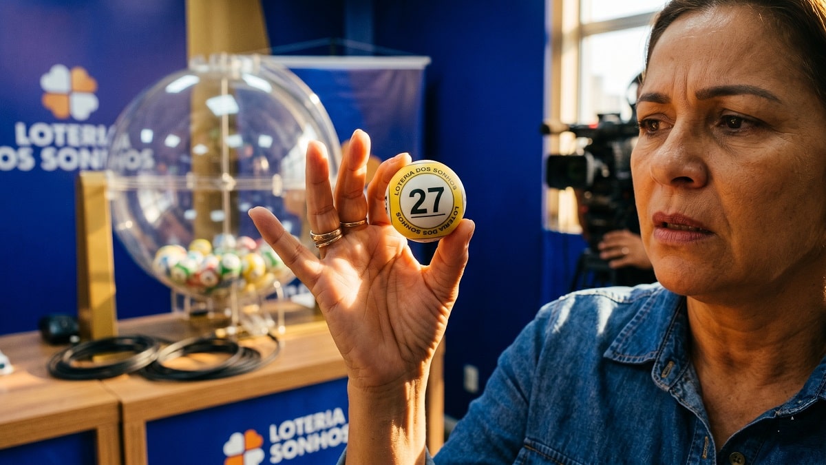 Mulher exibe bola número 27 da Loteria dos Sonhos durante sorteio, aparelho ao fundo