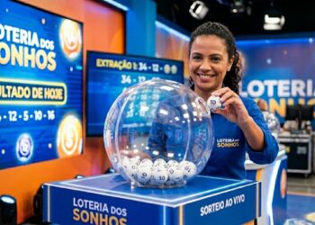 Mulher sorridente exibe bola com número 10 durante sorteio ao vivo da Loteria dos Sonhos, cenário azul e painéis digitais