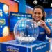 Mulher sorridente exibe bola com número 10 durante sorteio ao vivo da Loteria dos Sonhos, cenário azul e painéis digitais