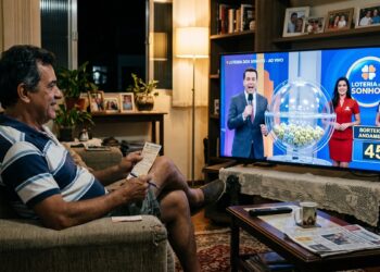 Homem sorridente confere bilhete enquanto assiste ao sorteio ao vivo da Loteria dos Sonhos na TV