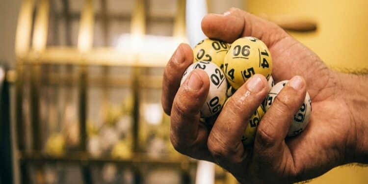 Mão masculina segurando bolas numeradas amarelas e brancas com números 02, 06, 14 e outros, globo ao fundo
