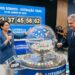 Apresentadora e homem em sorteio da Loteria dos Sonhos, atrás de globo com bolas numeradas, painel azul ao fundo