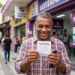 Homem sorridente de camisa xadrez amarela e roxa segura bilhete da Lotofácil 3700 e celular em frente à lotérica Caixa