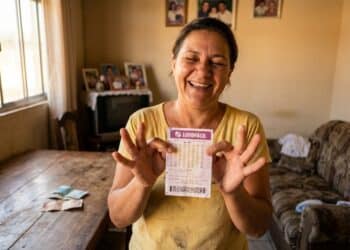 Mulher de meia-idade sorridente segura volante da Lotofácil em sala com fotos de família e TV ao fundo