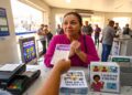 Mulher recebe bilhete da Lotofácil com expressão ansiosa em lotérica cheia, cartazes e panfletos visíveis