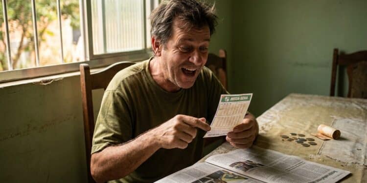 Homem sorridente aponta para bilhete da Mega-Sena ao lado de jornal e moedas na mesa