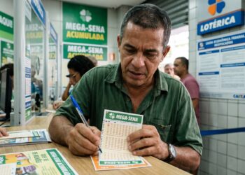 Homem de camisa verde marca números no volante da Mega-Sena dentro de lotérica, cartazes ao fundo destacam prêmio acumulado