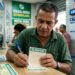 Homem de camisa verde marca números no volante da Mega-Sena dentro de lotérica, cartazes ao fundo destacam prêmio acumulado