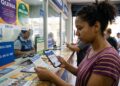 Mulher de blusa listrada roxa segura bilhete da Quina e celular em frente ao balcão de lotérica Caixa