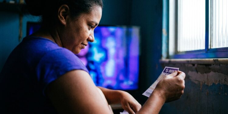 Mulher de camiseta azul segura bilhete da Quina em ambiente doméstico com TV ao fundo