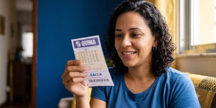 Mulher sorridente com cabelo cacheado segurando bilhete da Quina dentro de casa