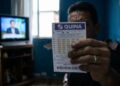 Homem exibe bilhete da Quina com TV ligada ao fundo em sala simples