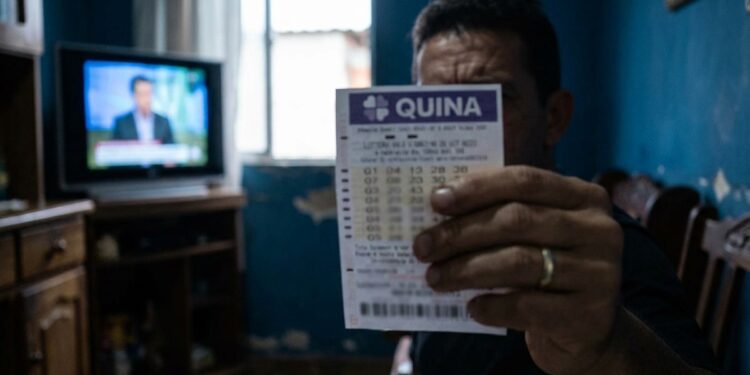 Homem exibe bilhete da Quina com TV ligada ao fundo em sala simples