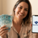Mulher sorridente segura notas de R$100 e celular com logo da Receita Federal em escritório.