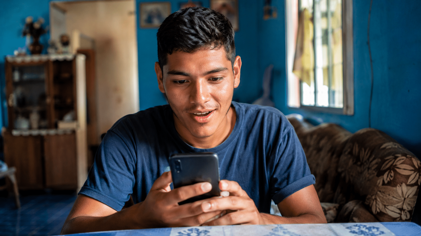 Rapaz de cabelo curto sentado à mesa olhando surpreso para o celular em ambiente doméstico azul.