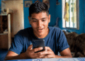 Rapaz de cabelo curto sentado à mesa olhando surpreso para o celular em ambiente doméstico azul.