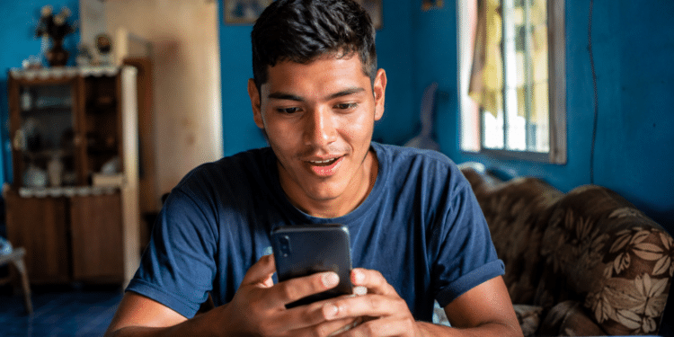 Rapaz de cabelo curto sentado à mesa olhando surpreso para o celular em ambiente doméstico azul.