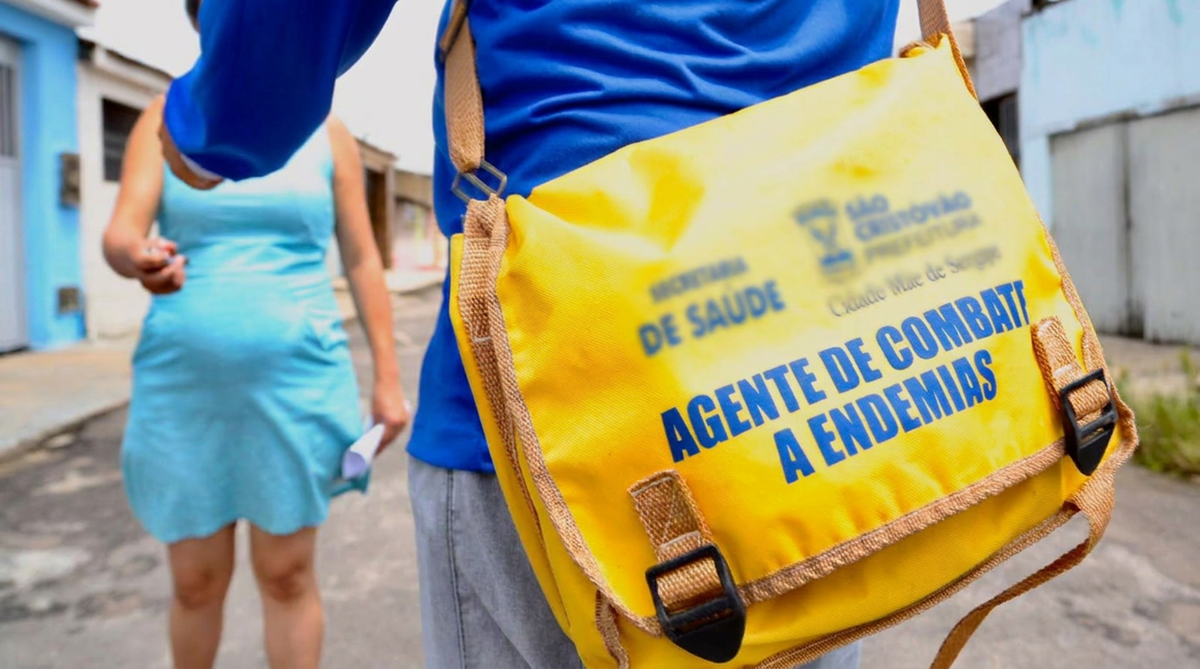 Agente de combate às endemias com bolsa amarela identificada faz visita em rua residencial