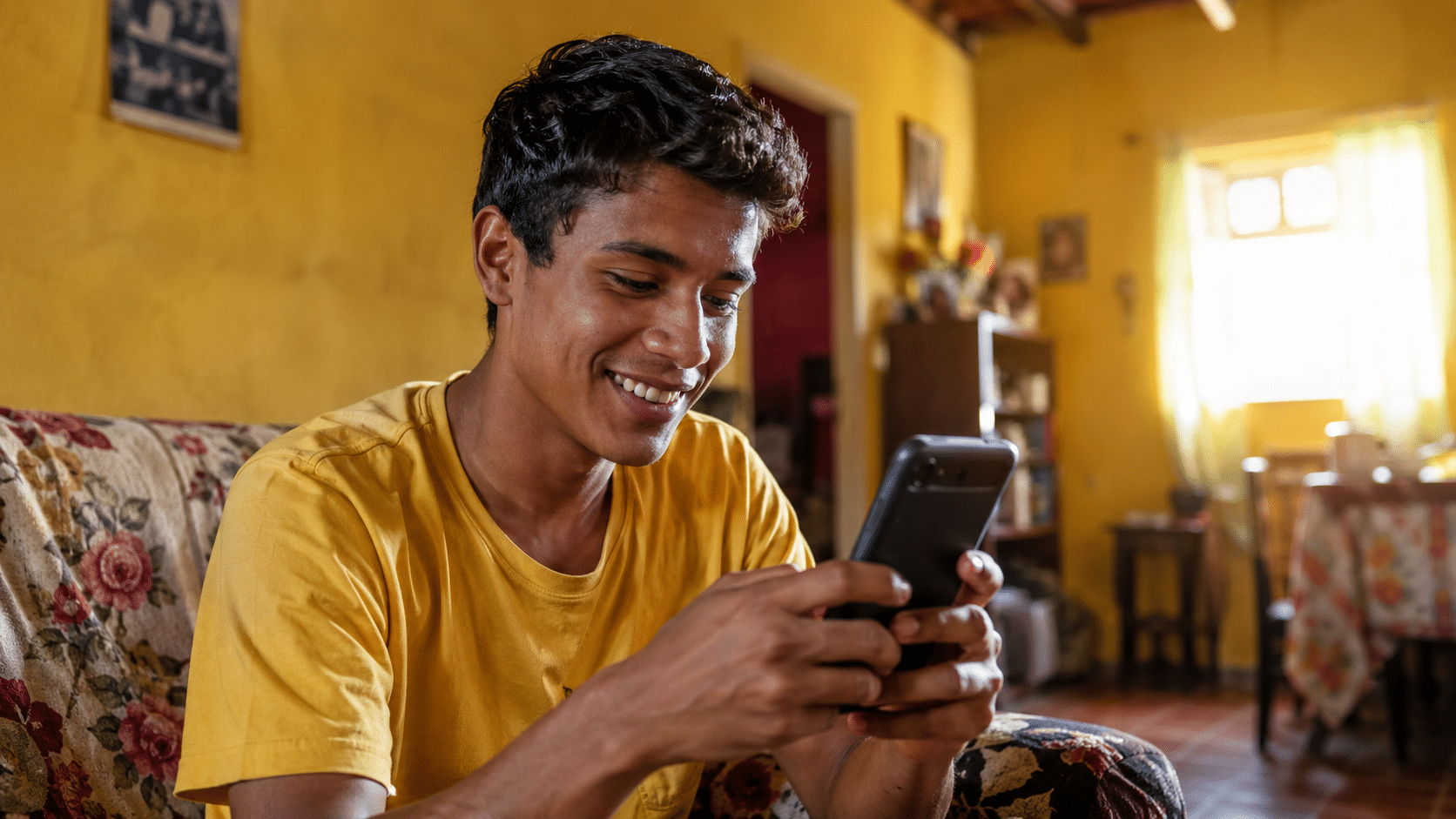 Jovem de cabelo curto sorri enquanto digita no celular sentado em sofá floral em sala iluminada.
