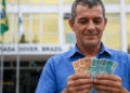 Servidor público sorri ao contar cédulas de 50 e 100 reais em frente a prédio governamental com bandeiras