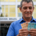 Servidor público sorri ao contar cédulas de 50 e 100 reais em frente a prédio governamental com bandeiras