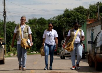 Três agentes de saúde caminham por uma rua com bolsas de trabalho a tiracolo durante visita de campo