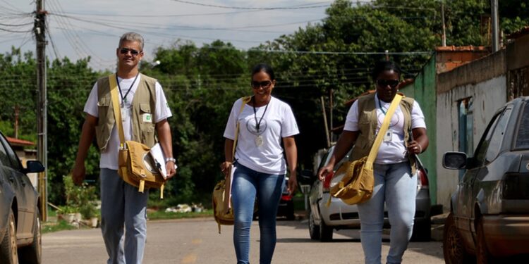 Três agentes de saúde caminham por uma rua com bolsas de trabalho a tiracolo durante visita de campo