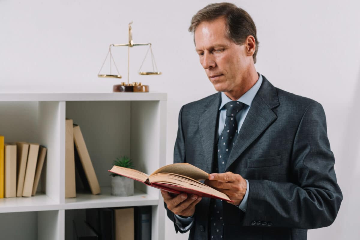 Advogado de terno e gravata lê um livro jurídico ao lado de uma balança da justiça e prateleira com livros