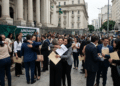Grupo de aprovados em concurso se abraça e comemora segurando documentos em frente a um prédio público