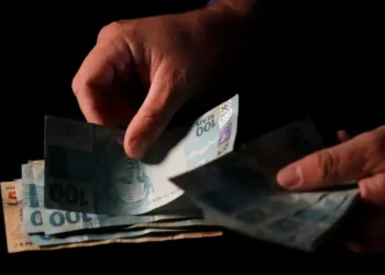 Mãos seguram notas de R$ 100 em imagem sobre pagamento do Auxílio Reconstrução pela Caixa.