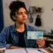 Estudante segura uma nota de R$ 100 em um quarto de estudos, com expressão de dúvida, representando o processo de geração do boleto para pagamento da taxa do Enem 2026 no último dia do prazo.