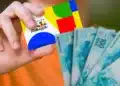 Mãos segurando cartão do Bolsa Família com cédulas de reais ao lado, representando pagamentos do benefício em julho.