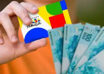 Mãos segurando cartão do Bolsa Família com cédulas de reais ao lado, representando pagamentos do benefício em julho.