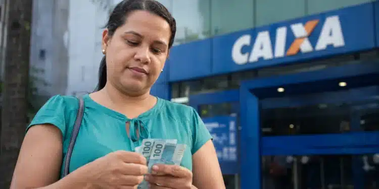 Mulher segura cédulas de reais em frente a uma agência da Caixa, representando os pagamentos do Bolsa Família de junho.
