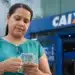 Mulher segura cédulas de reais em frente a uma agência da Caixa, representando os pagamentos do Bolsa Família de junho.