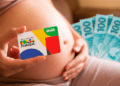 Gestante segura cartão do Programa Bolsa Família com logotipo colorido enquanto mostra notas de R$ 100 ao fundo
