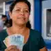 Mulher sorri segurando cédulas de reais diante de agência bancária azul, representando os pagamentos do Bolsa Família em junho.