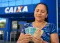 Mulher segura cédulas de reais em frente a uma agência da Caixa, representando o último repasse de junho do Bolsa Família.