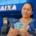 Mulher segura cédulas de reais em frente a uma agência da Caixa, representando o último repasse de junho do Bolsa Família.