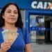 Mulher segurando cédulas de dinheiro em frente a uma agência da Caixa, representando depósitos de R$ 600, R$ 800 e R$ 1.000.