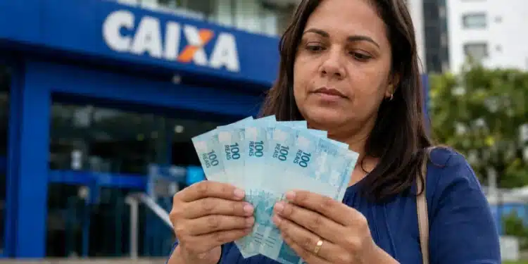 Mulher segurando cédulas de real em frente a agência da Caixa Econômica Federal, representando liberação de saque de R$ 6.220