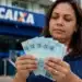 Mulher segurando cédulas de real em frente a agência da Caixa Econômica Federal, representando liberação de saque de R$ 6.220
