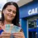 Mulher segura cédulas de dinheiro em frente a uma agência da Caixa, em referência à liberação de R$ 800 e R$ 1.000 no São João.