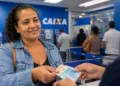 Mulher recebendo dinheiro em agência da Caixa, representando o pagamento de até R$ 1.000 para milhões de famílias brasileiras.